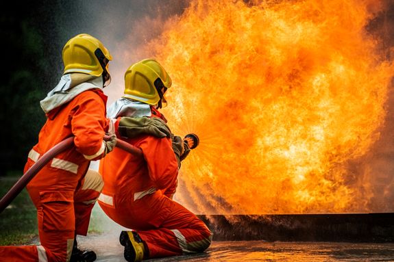 Two brave firefighter in fire suit on rescue duty using water from hose extinguishing fighting with big crackle fire flames inside burning premises (1)